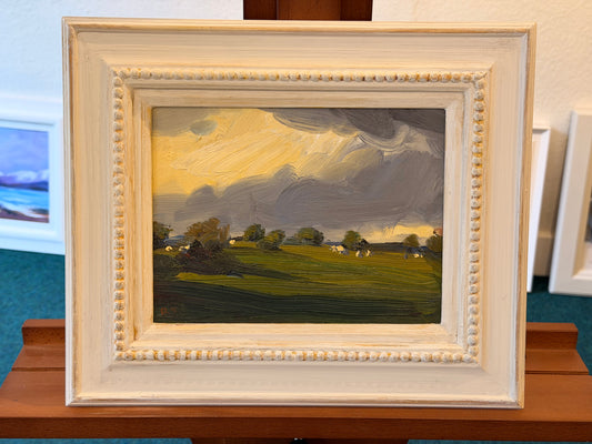 A framed landscape oil painting featuring a green field with trees and cows under a dramatic yellow and gray sky, housed in a distressed cream-colored wooden frame.