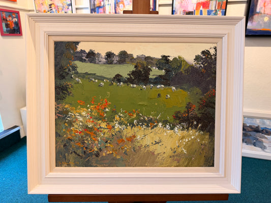 A framed landscape oil painting on an easel, featuring a green field with grazing sheep, trees, and wildflowers in the foreground. The painting is housed in a thick, white, decorative square frame.