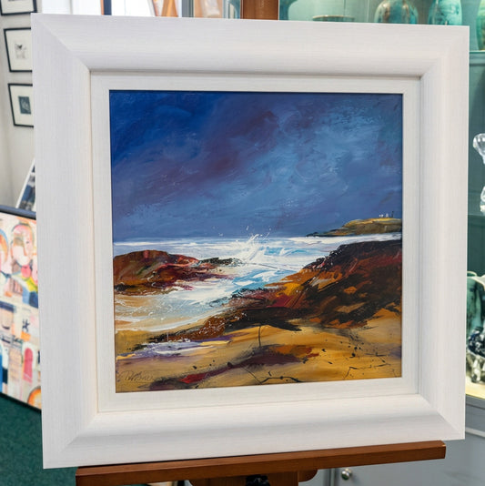 A framed landscape painting of a rocky beach and ocean waves, displayed on an easel in an indoor gallery or studio setting. The artwork features a dark blue sky, brown cliffs, and white surf, housed in a wide white frame.