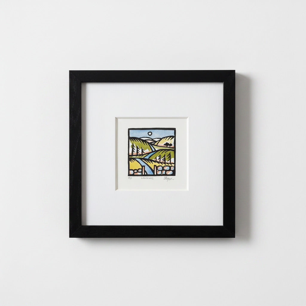 A framed art print featuring a colorful landscape illustration with green hills, a blue river, and a yellow sun, presented in a black frame with a white mat against a light gray wall.