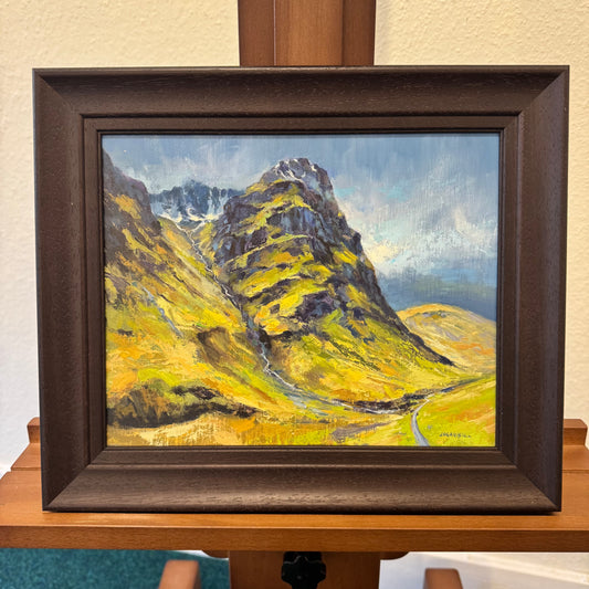 A framed landscape oil painting of a mountain peak with yellow-green slopes, a winding road, and a blue sky, displayed on a wooden easel.