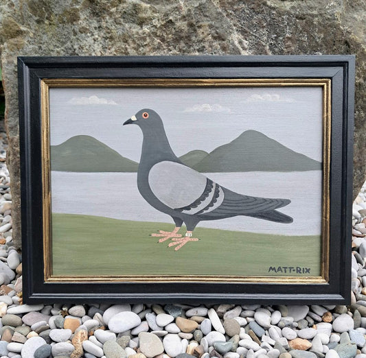 Framed painting of a pigeon with a landscape background, placed on pebbles.