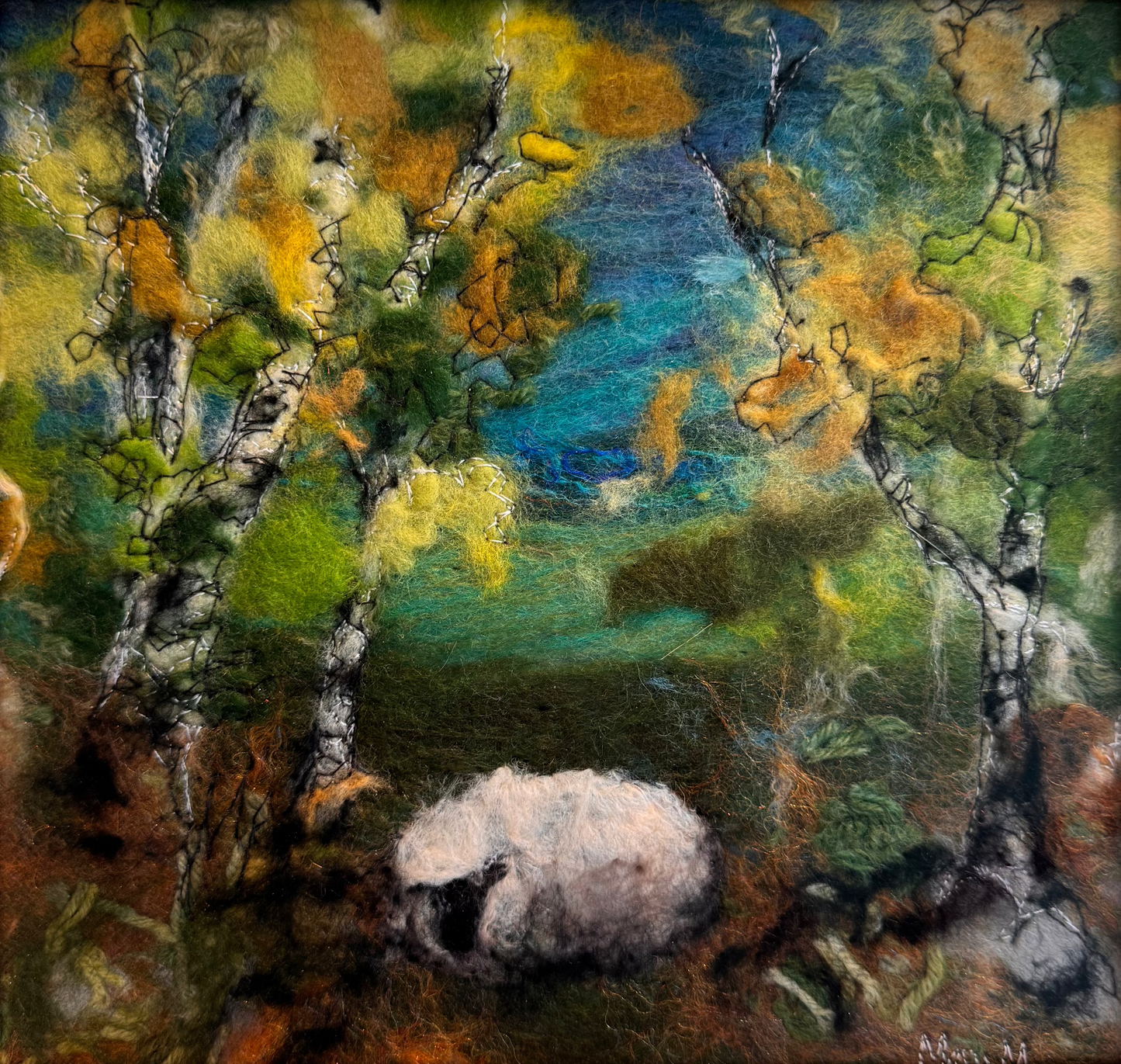 A framed felt painting depicting a white sheep grazing among birch trees with green, yellow, and blue foliage, set in a light wood frame.