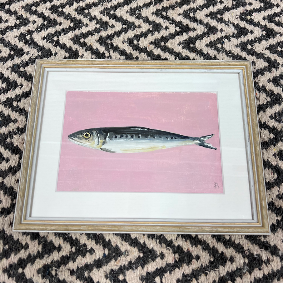 Framed artwork of a fish on a pink background, placed on a patterned surface.