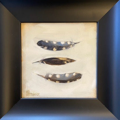 Framed wall art featuring three painted feathers on a cream background, set in a wide black frame and mounted on a light wall.