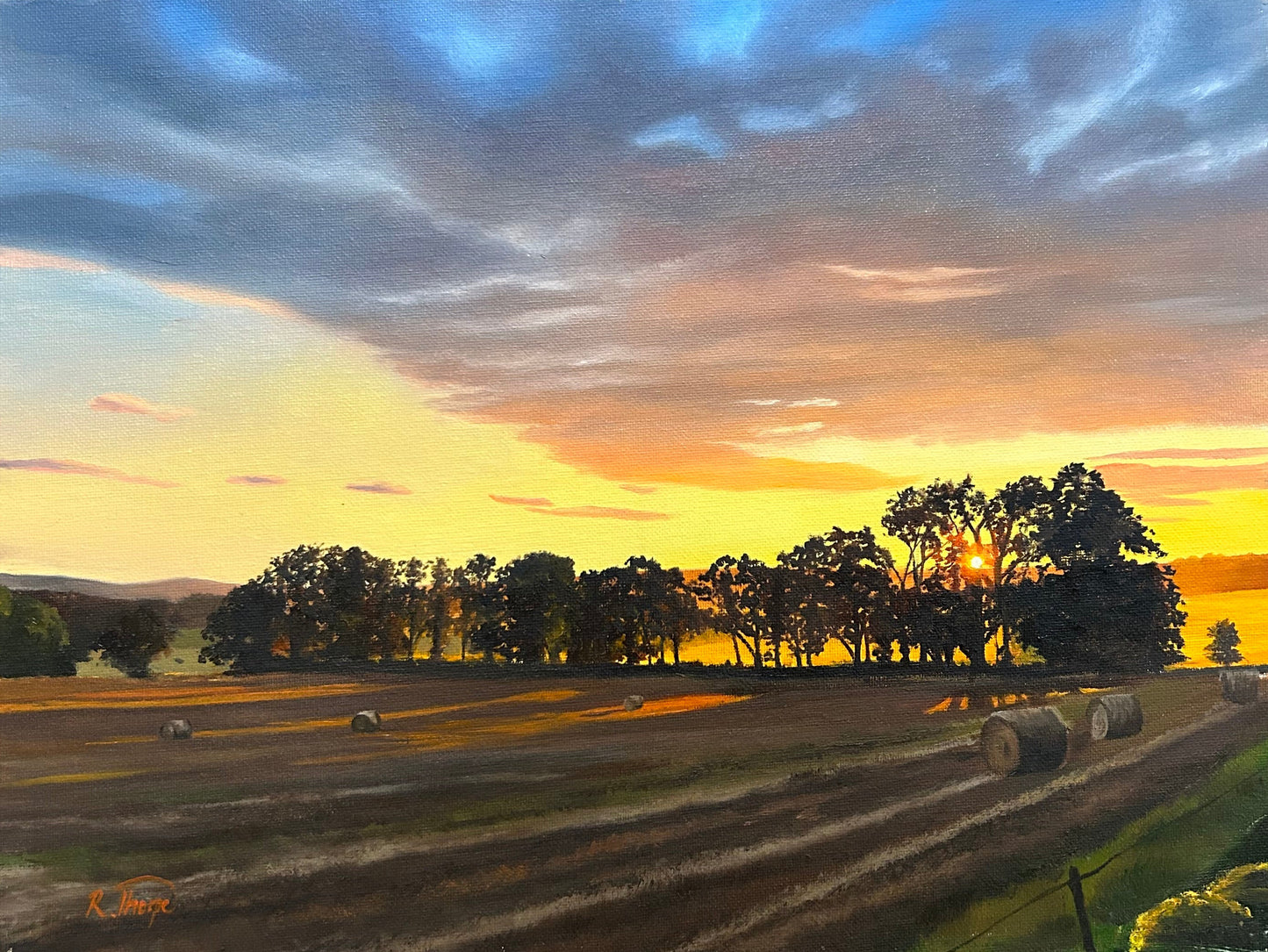 A landscape painting of a sunset over a harvested field with hay bales and trees, displayed against a dark wooden background.