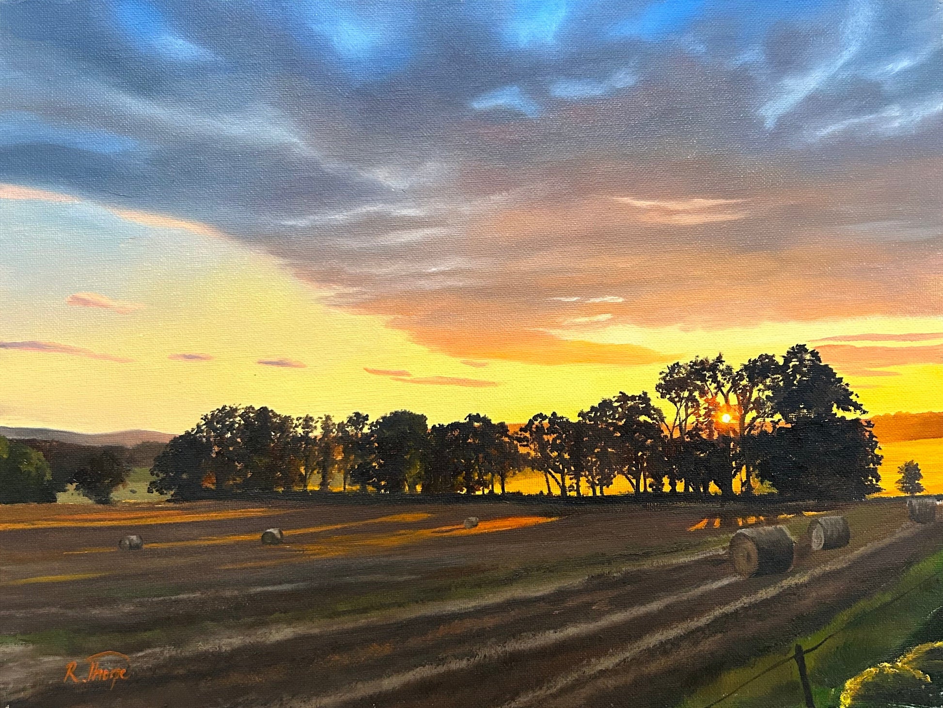 A landscape painting of a sunset over a harvested field with hay bales and trees, displayed against a dark wooden background.
