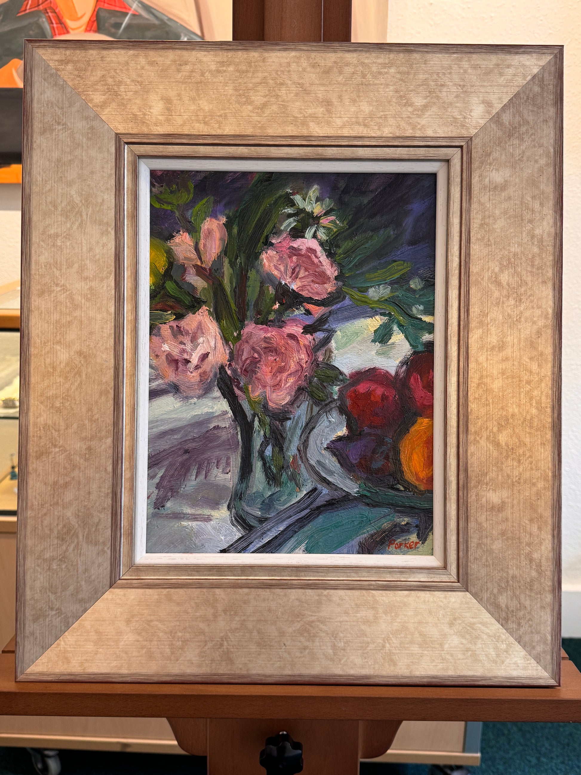 A framed still life painting featuring pink flowers in a vase and red and orange fruits, housed in a wide textured beige frame and displayed on a wooden easel.