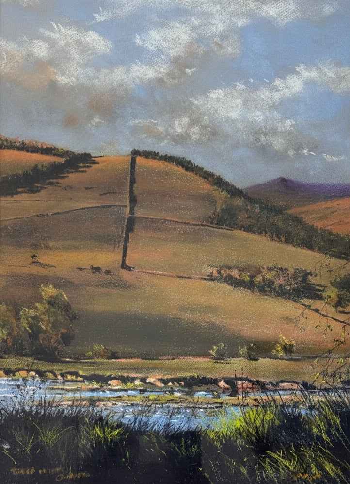 A framed landscape painting depicting rolling hills, a river in the foreground, and a cloudy sky, mounted on a light beige wall.