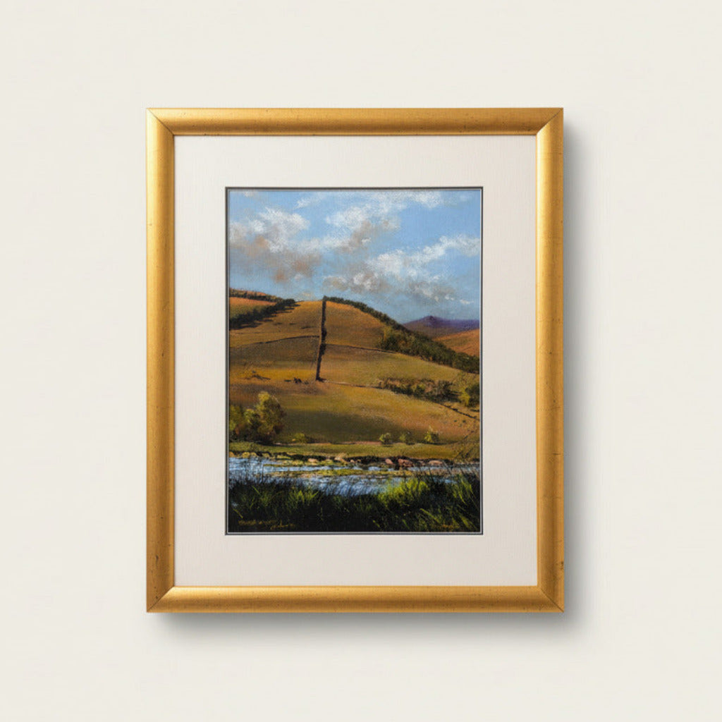A framed landscape art print featuring rolling hills, trees, and a river under a cloudy sky, mounted with a cream mat and a gold frame on a textured white wall.