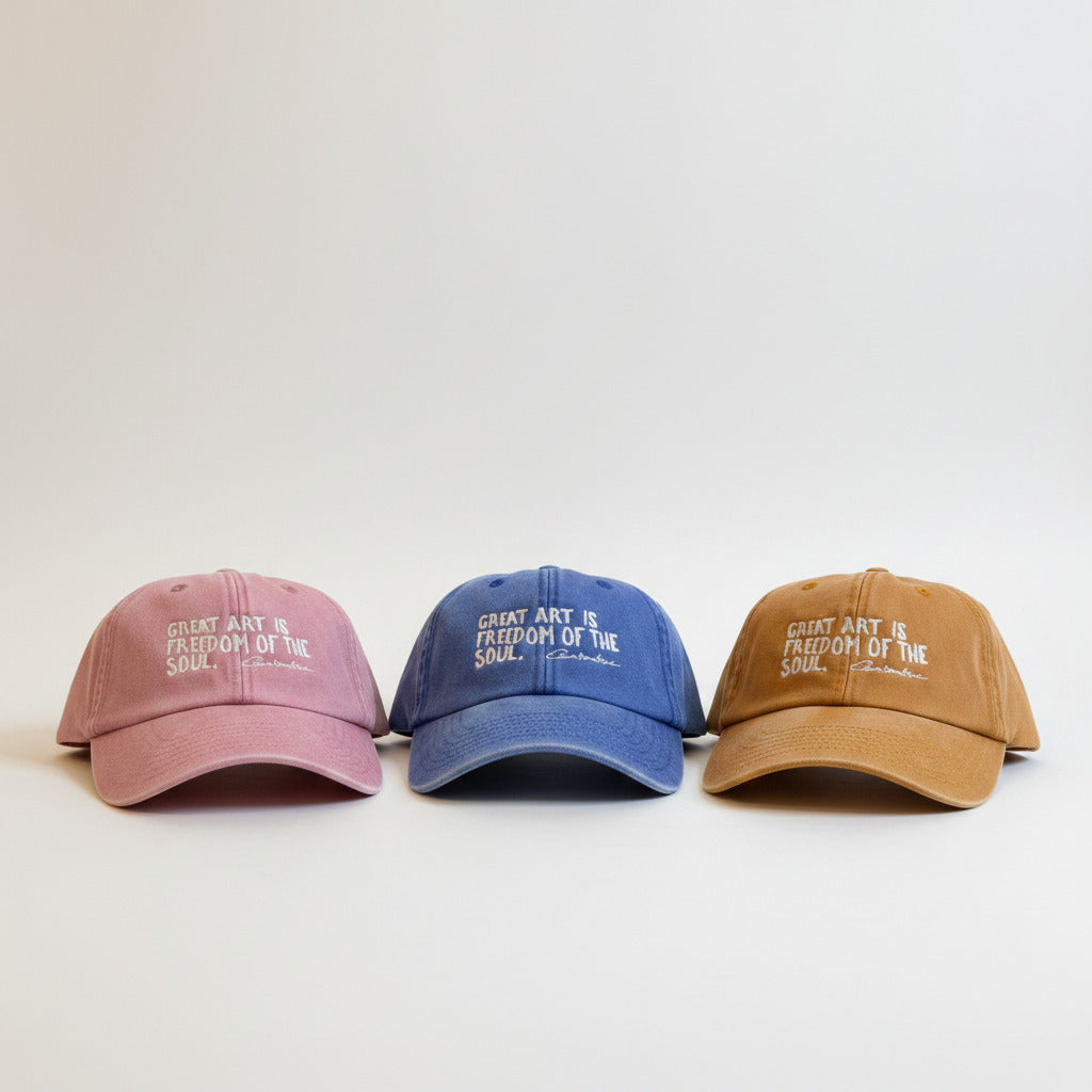 Three baseball caps in dusty pink, mustard yellow, and royal blue, each embroidered with the phrase 'GREAT ART IS FREEDOM OF THE SOUL.' in white lettering, arranged on a white surface against a textured beige wall.