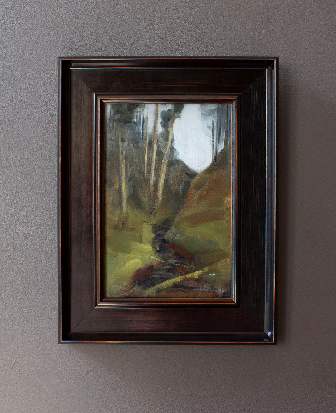 A framed landscape oil painting hanging on a textured gray wall. The painting depicts a forest scene with tall trees, a winding river, and rolling hills in earthy tones of green, brown, and yellow.