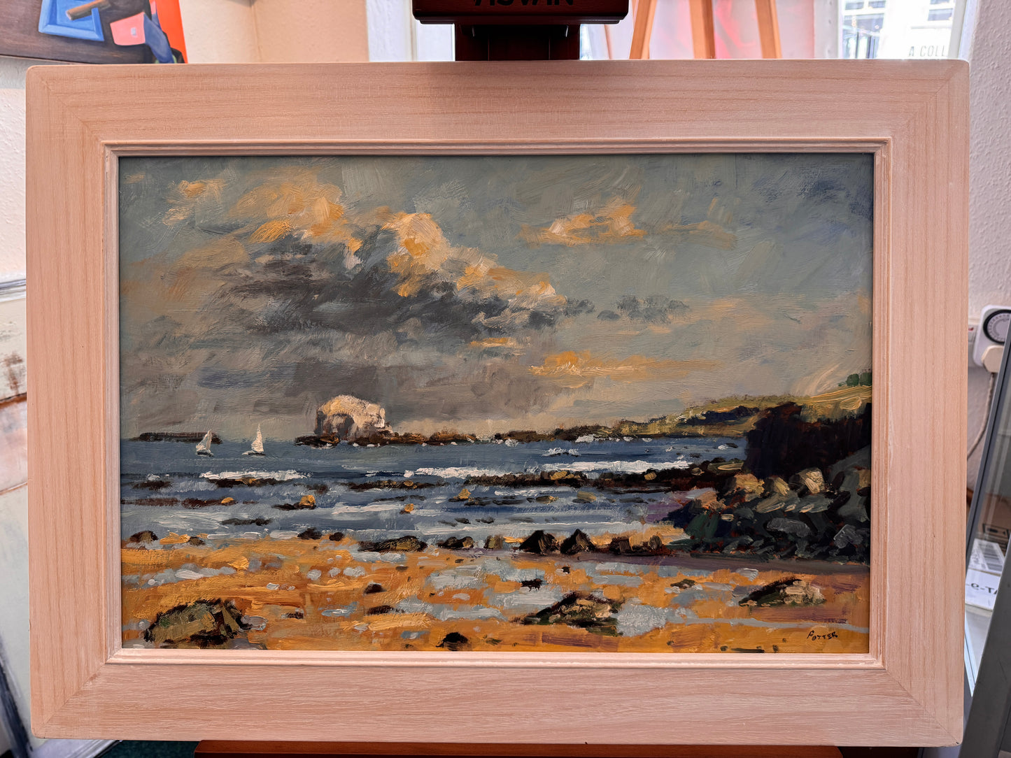 A framed oil painting of a coastal landscape with rocky shore, sea, and dramatic clouds, displayed on an easel. The painting features a sandy beach, dark rocks, sailboats, and a stone structure on the horizon under a cloudy sky.