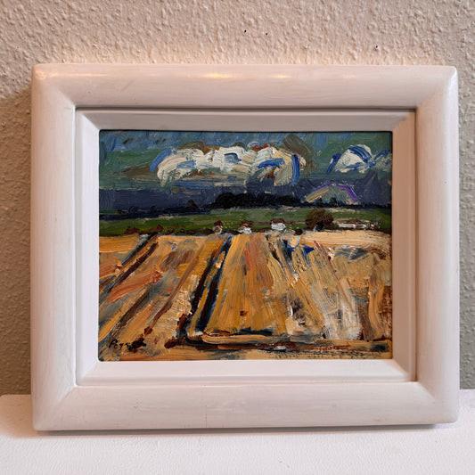 A framed landscape oil painting featuring a golden-brown field under a dramatic cloudy sky, housed in a white wooden frame and displayed against a textured beige wall.
