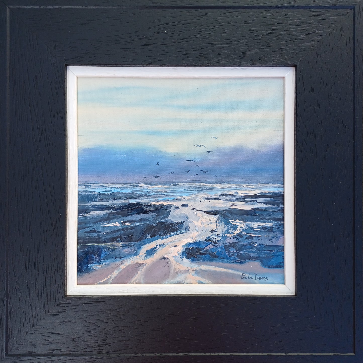 A framed coastal landscape painting featuring blue and white ocean waves, dark rocky shoreline, and a pale sky with small birds, presented in a dark textured frame with white matting.