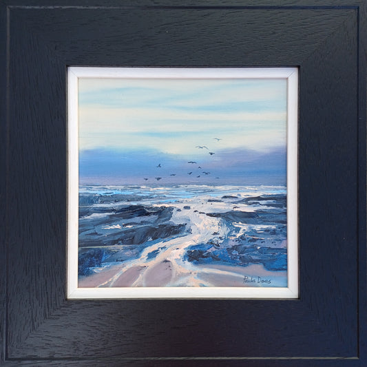 A framed coastal landscape painting featuring blue and white ocean waves, dark rocky shoreline, and a pale sky with small birds, presented in a dark textured frame with white matting.