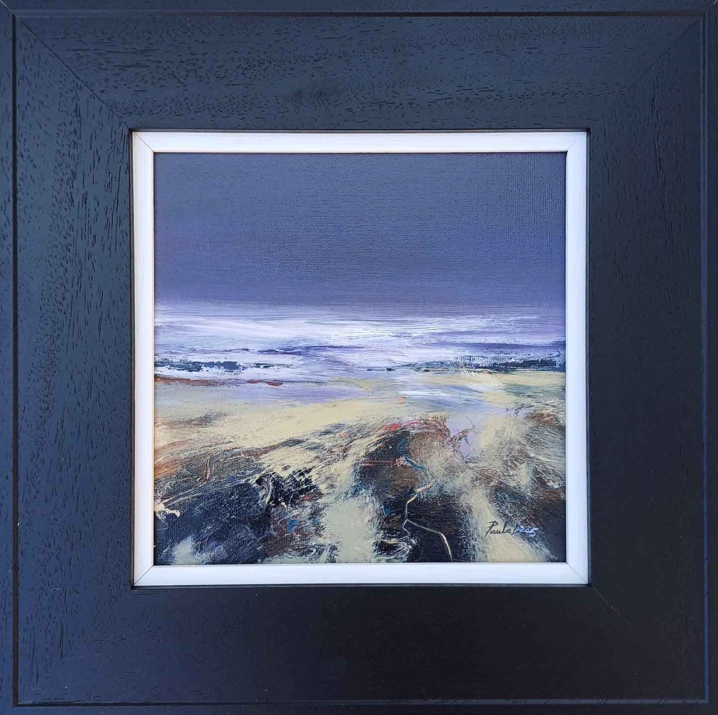 Framed landscape painting featuring a beach scene with a dark blue-gray sky, white surf, and sandy shore in textured brushstrokes, set within a dark textured frame with a white mat.