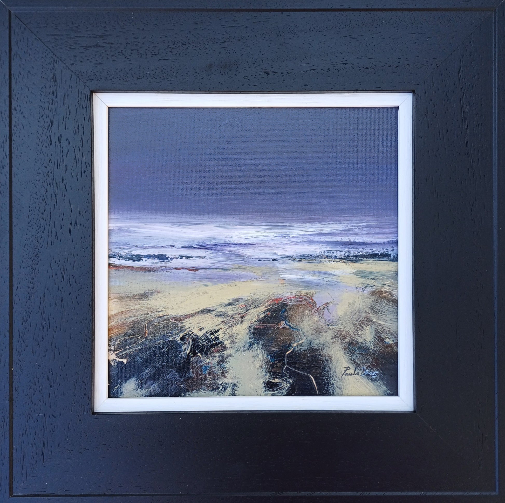 Framed landscape painting featuring a beach scene with a dark blue-gray sky, white surf, and sandy shore in textured brushstrokes, set within a dark textured frame with a white mat.