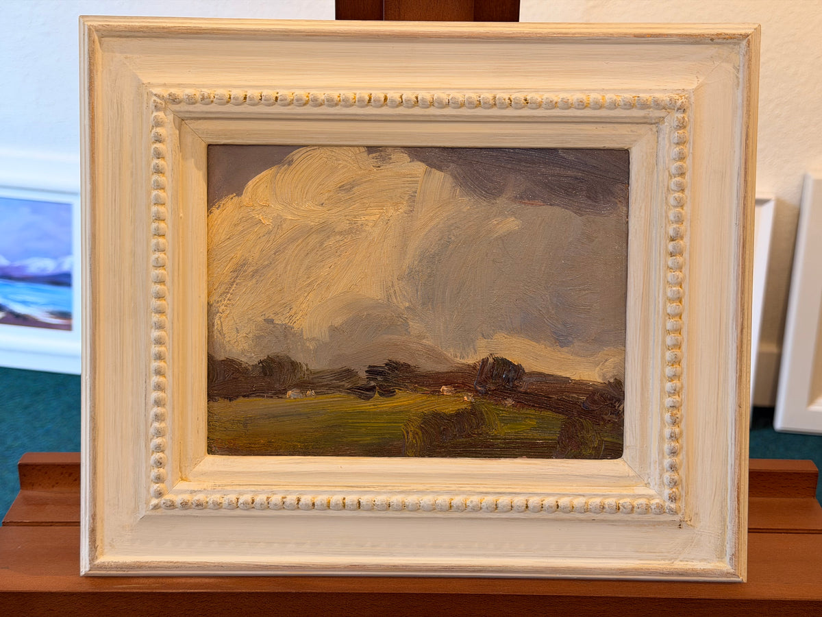 A framed landscape oil painting with a dramatic cloudy sky, shown in a distressed off-white wooden frame on a wooden display stand.