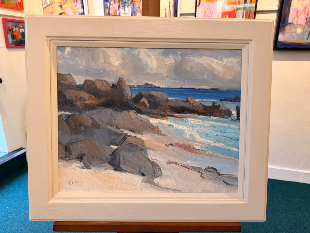 A framed coastal landscape painting displayed on an easel in an art gallery or studio. The painting depicts a rocky beach with blue ocean water, white surf, and a cloudy sky, set within a wide cream-colored frame.