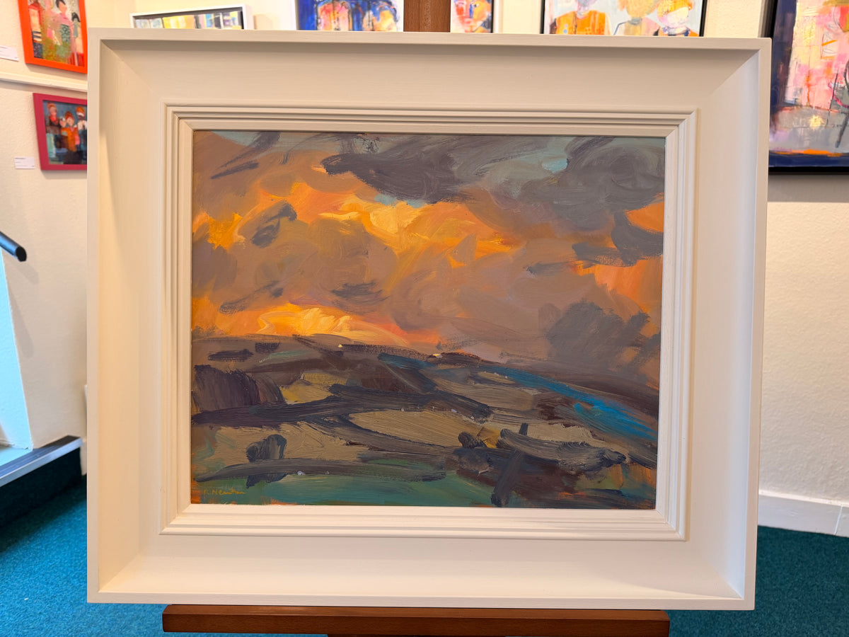 A framed landscape oil painting on an easel, featuring a dramatic sunset sky in orange, yellow, and gray with dark clouds over a coastal or rocky terrain. The painting is housed in a thick, white square frame.