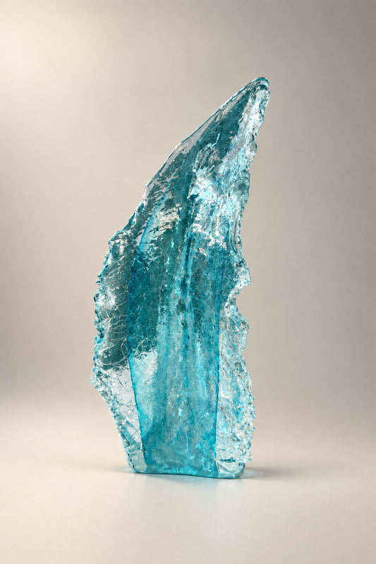 A tall, translucent light blue glass sculpture standing on a white pedestal in an indoor gallery or studio space with framed artwork on the wall behind it.