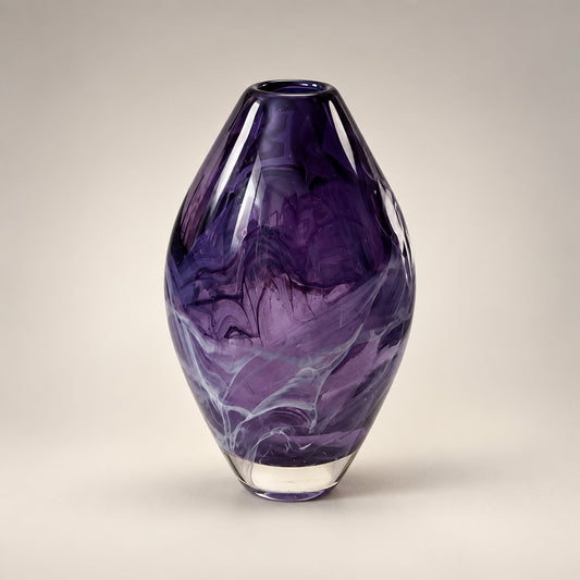 A purple glass vase with a rounded, bulbous shape and a flared base, sitting on a white table in an indoor room with green carpet and framed landscape paintings in the background.