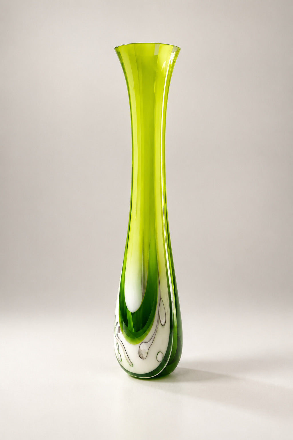 A tall, flared glass vase with a vibrant lime green and dark green gradient and a white swirl pattern near the base, displayed on a white table in an art gallery.