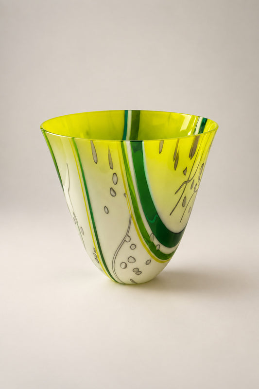 A hand-blown glass bowl with a vibrant yellow and white swirl pattern and green accents, displayed on a white pedestal in an indoor setting.