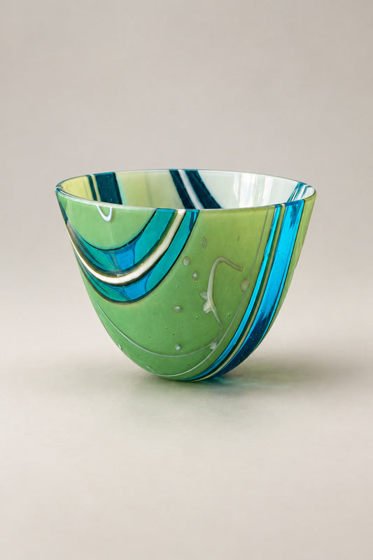 A hand-blown glass bowl with green, blue, and white swirls, sitting on a polished green marble table in an indoor setting.