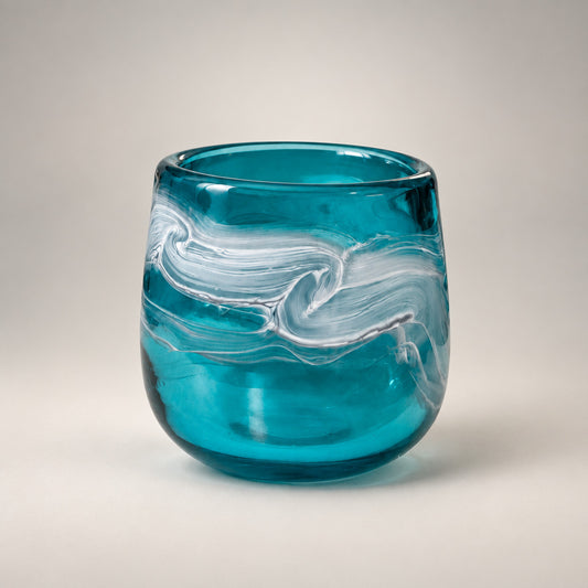 A small round glass tumbler with a swirled teal blue and white pattern, sitting on a white tabletop in an indoor setting.