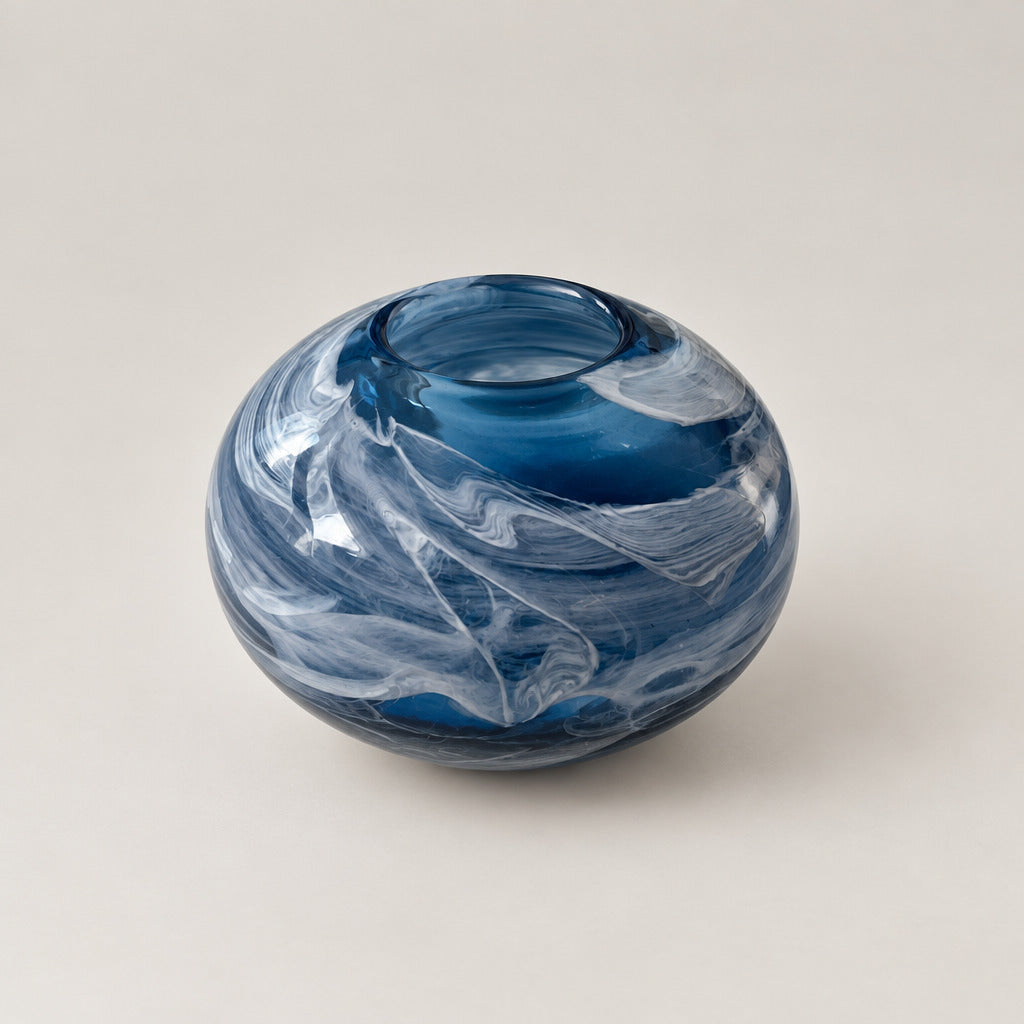 A small round glass vase with a marbled swirl pattern in deep blue and white, sitting on a white surface in an indoor setting.