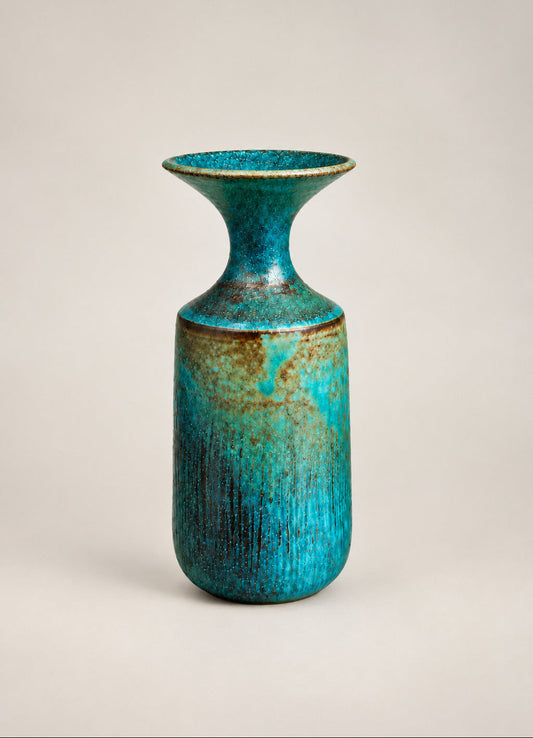 A handmade ceramic vase with a flared neck and a textured, distressed glaze in shades of teal, blue, and brown, displayed on a glass shelf against a textured off-white wall.