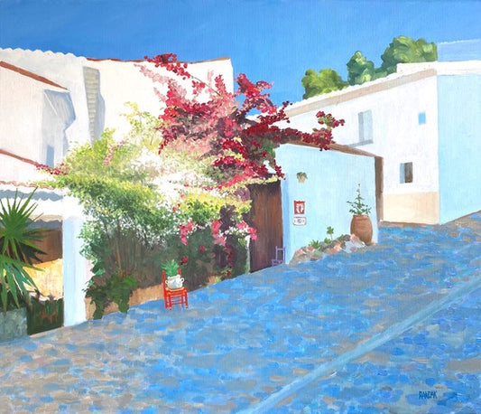 Cobbled street with white buildings, red chair, and flowering plants under a blue sky.
