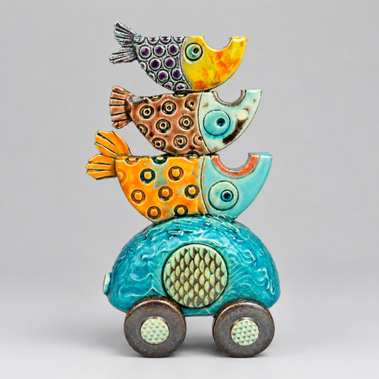 A colorful ceramic sculpture of three stacked fish riding on top of a turquoise car, photographed against a dark blue-to-gray gradient studio background.