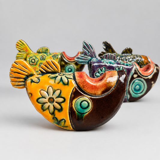 A small fish-shaped ceramic ornament with colorful floral and swirl patterns, arranged on a light surface against a dark blue background.