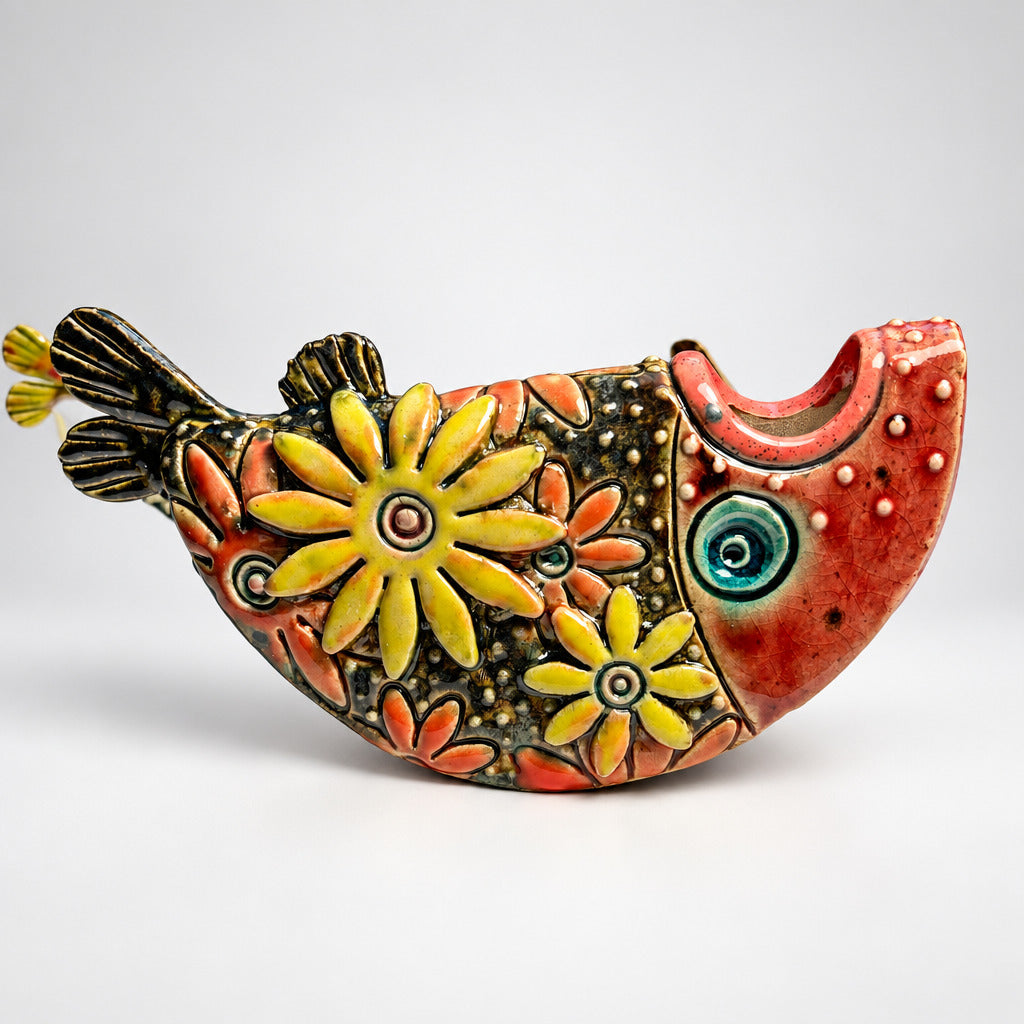 A handcrafted ceramic fish-shaped object with a coral-pink body, yellow and orange floral patterns, and a blue-green eye, photographed against a dark blue to light gray gradient background.