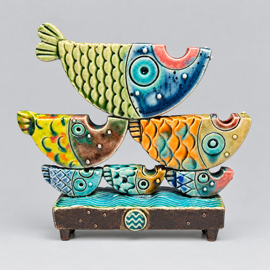 A colorful ceramic fish sculpture set on a dark base, featuring stacked stylized fish in green, blue, yellow, and orange with glossy painted glazes against a dark studio background.