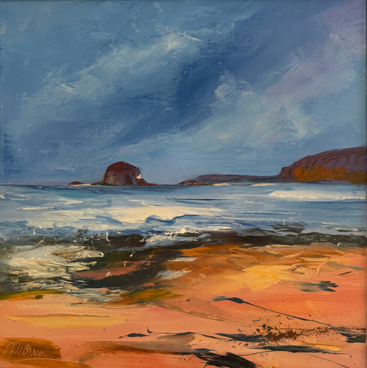 A framed landscape painting featuring a beach scene with blue sky, water, and warm orange-brown sand, housed in a light wood frame. A faint reflection of a person is visible on the glass.
