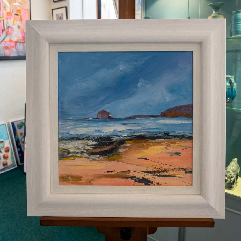 A framed landscape painting of a rocky beach and sea under a blue sky, displayed on a wooden easel in an art studio. The frame is white, and the painting features blue, pink, and brown tones.