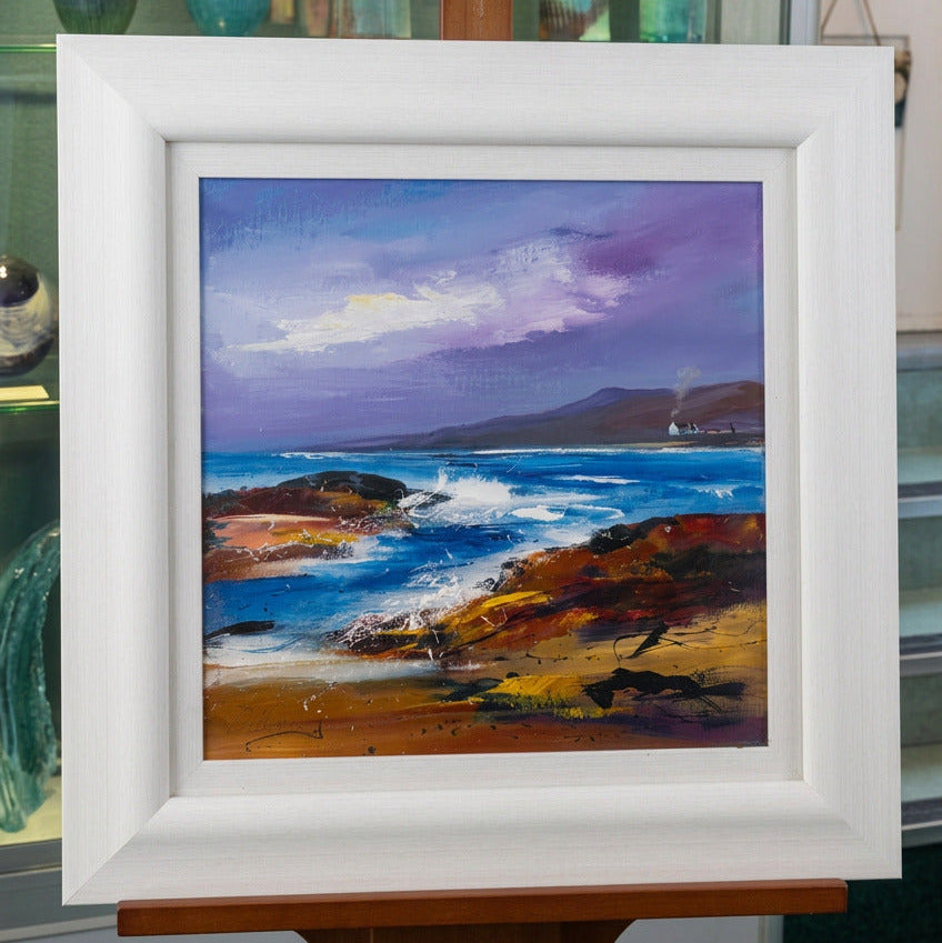 A framed coastal landscape painting displayed on an easel. The artwork depicts a dramatic purple and blue sky over rocky shoreline waves, with a light wood frame around the print.