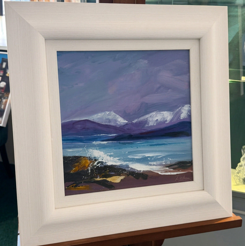 A framed landscape painting of snowy mountains, a lake, and a beach, displayed on a wooden easel in a gallery or shop. The frame is a light cream/off-white wood, and the painting features purple, blue, and white tones.