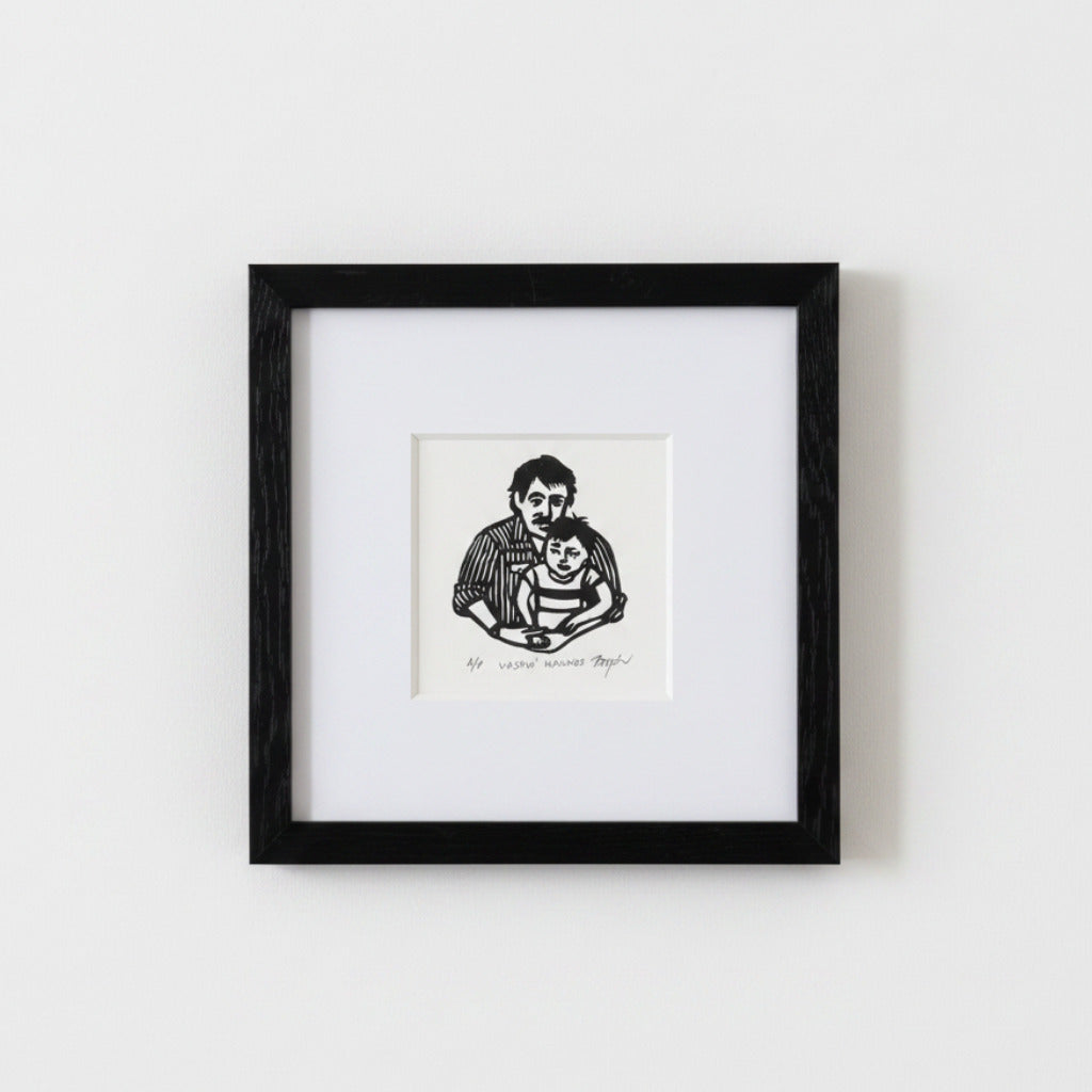A framed black and white art print depicting a father holding a child, mounted on a white mat within a black square frame against a light gray wall.