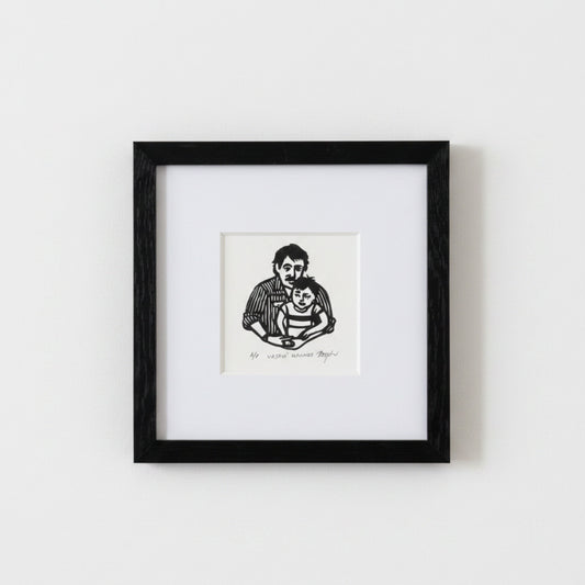 A framed black and white art print depicting a father holding a child, mounted on a white mat within a black square frame against a light gray wall.