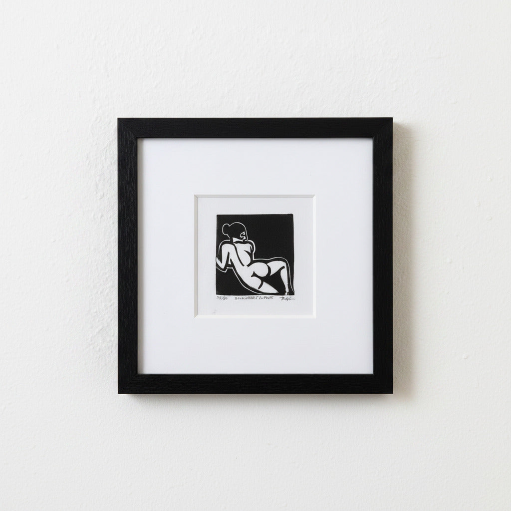 A framed black and white art print featuring a minimalist line drawing of a reclining nude figure, presented in a black frame with a white mat against a textured white wall.