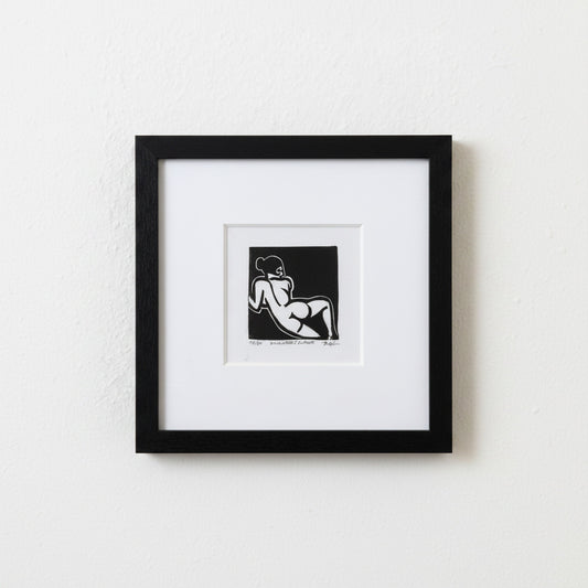 A framed black and white art print featuring a minimalist line drawing of a reclining nude figure, presented in a black frame with a white mat against a textured white wall.