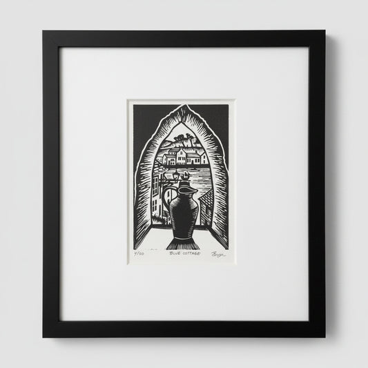 A framed black and white art print featuring a block-print style illustration of a house seen through an arched doorway with a large vase in the foreground. The print is matted in white and enclosed in a black frame, shown against a light gray wall.