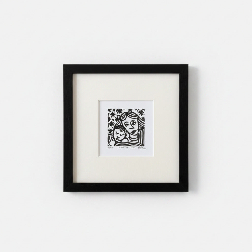 A framed black and white art print featuring a stylized illustration of a woman holding a child, mounted with a white mat inside a black square frame against a light gray wall.