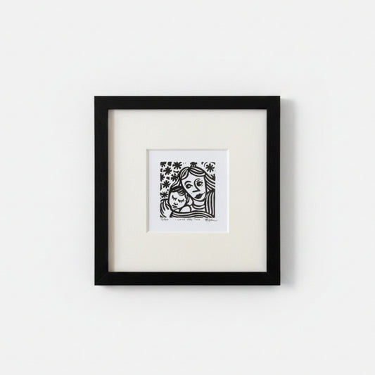 A framed black and white art print featuring a stylized illustration of a woman holding a child, mounted with a white mat inside a black square frame against a light gray wall.