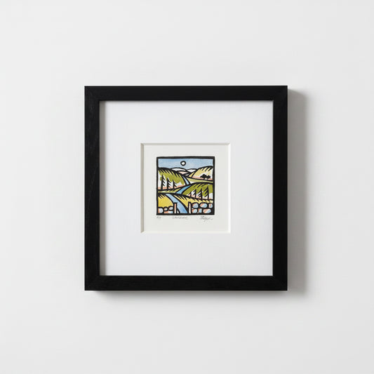 A framed art print featuring a colorful landscape illustration with green hills, a blue river, and a yellow sun, presented in a black frame with a white mat against a light gray wall.